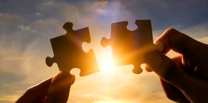 Two hands holding puzzle pieces in front of the sun.